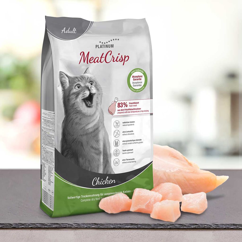 Vrečka PLATINUM MeatCrisp Adult Chicken, Suha Hrana za Mačke stoji pokonci ob surovem piščancu na mizi. Na embalaži je prikazana srečna siva mačka, podrobnosti o izdelku in poudarjen postopek Freshmeatdryer.