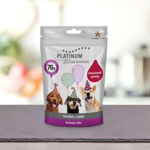 Vrečka PLATINUM Click-Bits Chicken+Lamb - zdrava pasja nagrada, na kateri so trije psi v party klobučkih z baloni v ozadju.