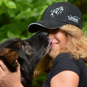 Nasmejano osebo v Ekskluzivna PLATINUM kapa s šiltom blagovne znamke PLATINUM po obrazu nežno oblizuje velik črno-rjav pes, v ozadju pa je zeleno listje.