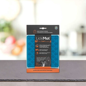 Modra PLATINUM Podloga LickiMat® - TUFF BUDDY v embalaži stoji pokonci na sivi površini z zamegljenim notranjim ozadjem. Na embalaži so navedene prednosti, kot je zmanjšanje tesnobe psa za pse in mačke.