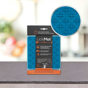 A PLATINUM Podloga LickiMat® - TUFF BUDDY v modri barvi v embalaži leži na mizi. Embalaža izpostavlja prednosti za hranjenje, treniranje in zmanjšanje tesnobe psa. Približan posnetek prikaže teksturiran križni vzorec površine podloge.