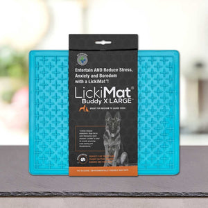 Modra podloga LickiMat® - BUDDY by PLATINUM za srednje velike in velike pse je prikazana na pultu, na embalaži pa so poudarjene njene lastnosti za zmanjševanje stresa in dolgočasja, ki spodbujajo počasno hranjenje psa.