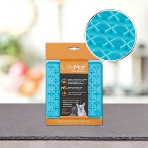Modra Podloga LickiMat® - BUDDY by PLATINUM v oranžni embalaži stoji na mizi in prikazuje informacije o izdelku in sliko psa. Vložek poudarja njeno teksturirano površino za počasno hranjenje psa.