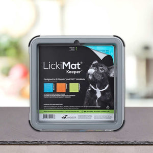 Na mizi stoji škatla PLATINUM LickiMat® - BUDDY KEEPER, na embalaži je črn francoski buldog z vidnimi informacijami o izdelku in blagovno znamko. Mehko zamegljeno ozadje poudarja to praktično držalo za LickiMat podloga za lizanje.