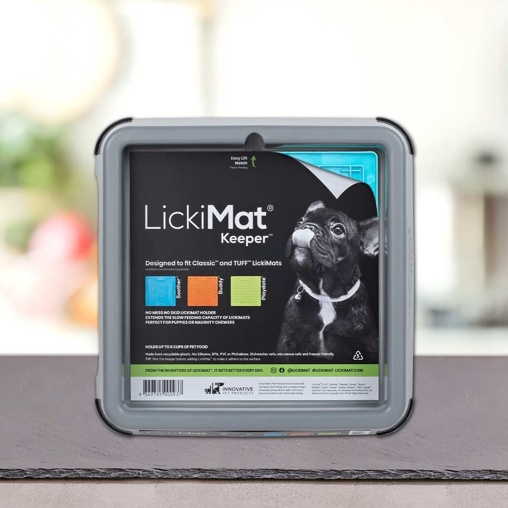 Na mizi stoji škatla PLATINUM LickiMat® - BUDDY KEEPER, na embalaži je črn francoski buldog z vidnimi informacijami o izdelku in blagovno znamko. Mehko zamegljeno ozadje poudarja to praktično držalo za LickiMat podloga za lizanje.