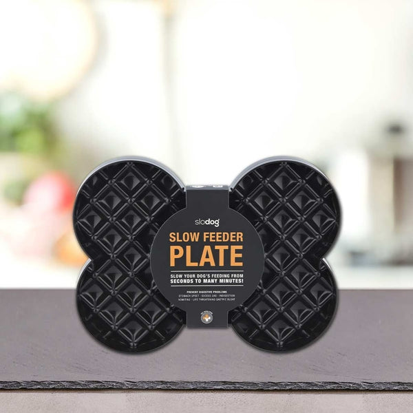Črna podloga LickiMat® v obliki kosti - SLODOG by PLATINUM stoji na pultu. Na embalaži je poudarjena njena uporaba za počasno hranjenje psov, za njo pa je zamegljeno notranje ozadje.