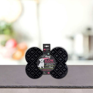 Črna plošča za počasno hranjenje v obliki kosti z napisom "Podloga LickiMat® - SLODOG" blagovne znamke PLATINUM, prikazana pokonci na kuhinjskem pultu z zamegljenim ozadjem; idealna za počasno hranjenje psov kot LickiMat ali pasja podloga za lizanje.