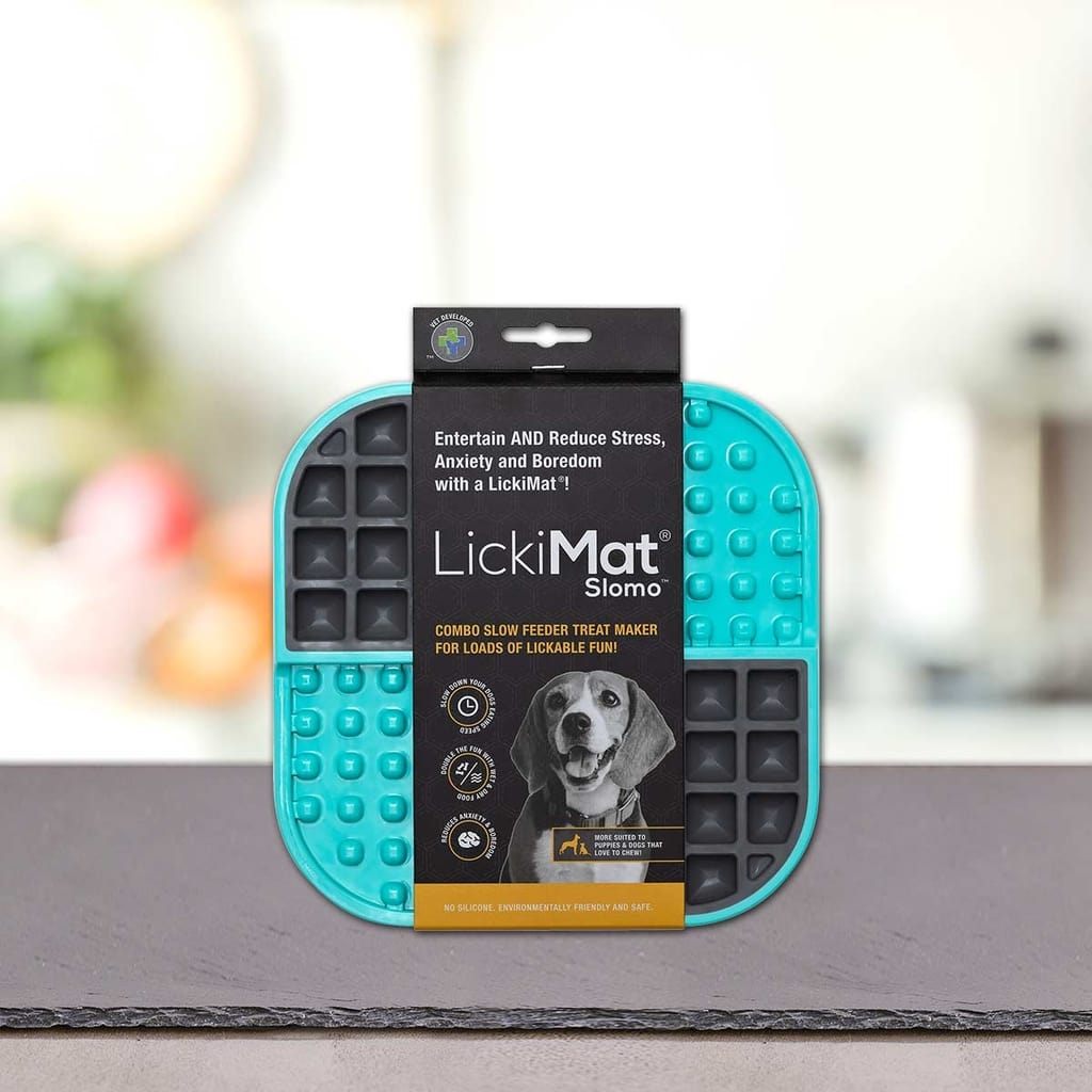 PLATINUM Podloga LickiMat® - SLOMO v modri in črni barvi je prikazana pokončno na sivi površini, na embalaži pa je ilustracija psa in poudarjene prednosti počasnega hranjenja. Mehko zamegljeno ozadje povečuje osredotočenost na izdelek.