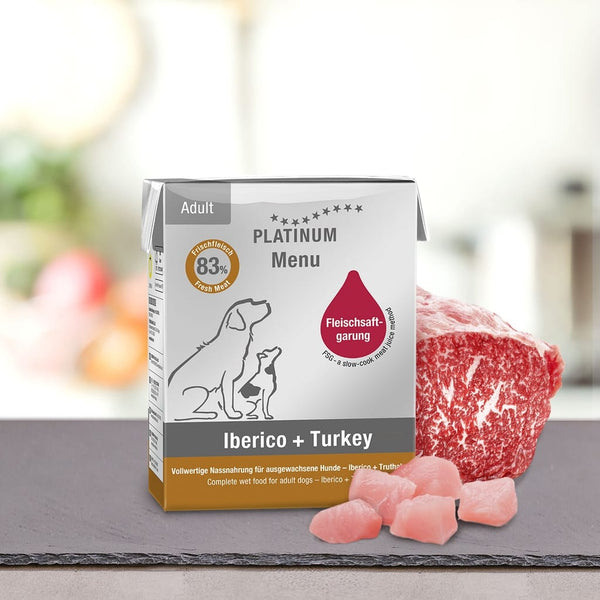 Škatla PLATINUM Poskusni paket različnih okusov Mokre Hrane za Pse je razstavljena poleg sveže meso za pse na pultu z zamegljenim zelenim ozadjem, na embalaži pa so ilustracije dveh psov.
