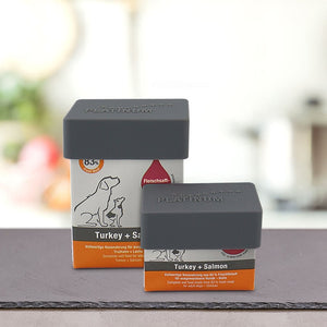 Dve posodi hrane za hišne ljubljenčke PLATINUM Turkey + Salmon s Praktičen Pokrovček MENU za shranjevanje Mokre Hrane proizvajalca PLATINUM ležita na sivi površini z ilustracijami psov in mačk. Nežno zamegljeno ozadje dodaja eleganco.