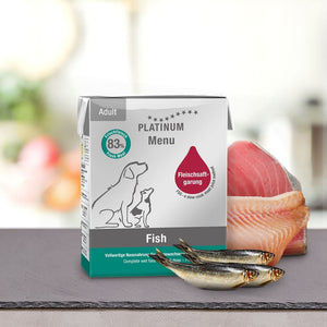 Na pultu je prikazana škatla PLATINUM Paket različnih okusov Mokre Hrane za Pse s sveže meso, koščki surovih rib, in dvema majhnima celima ribama spredaj, da se poudari visoka prebavljivost.