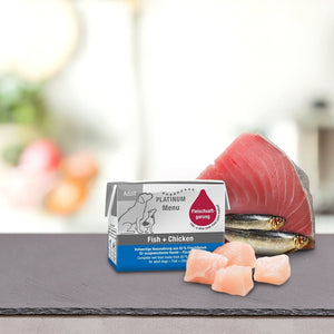 Škatla PLATINUM MENU Mini mokra hrana & DoggyCoach - Nagrada za tvojega psa je razstavljena na pultu s surovim ribjim filejem, sardinami in koščki piščanca na zamegljenem kuhinjskem ozadju.